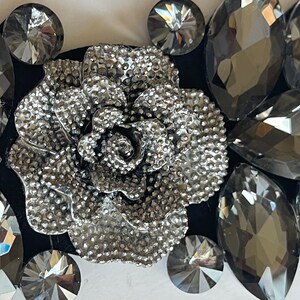 May include: A silver-toned metal rose with a textured surface, surrounded by black and gray rhinestones. The rose is a decorative element, possibly for a hair clip or a brooch.