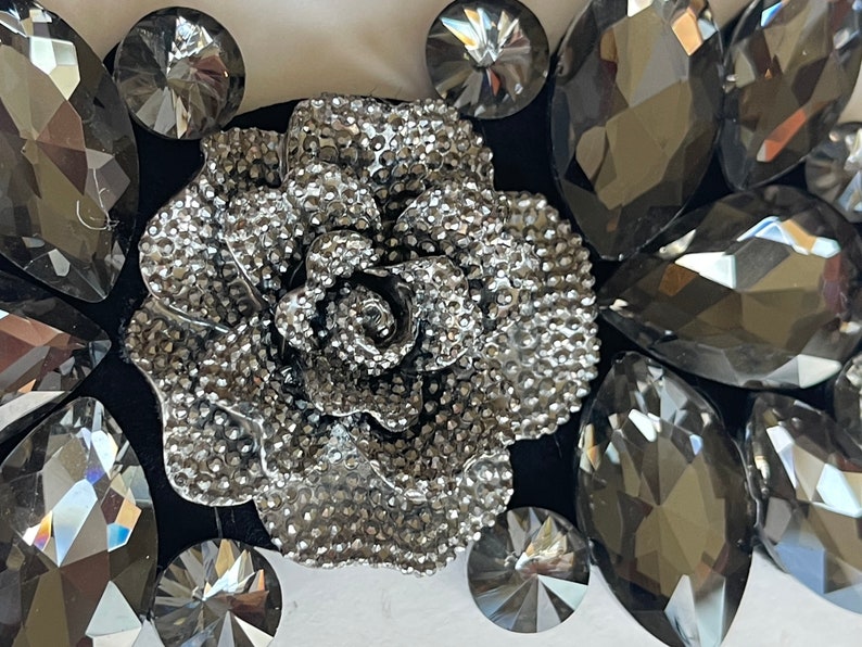 May include: A silver-toned metal rose with a textured surface, surrounded by black and gray rhinestones. The rose is a decorative element, possibly for a hair clip or a brooch.
