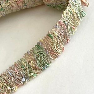 May include: A close-up of a multi-colored fringe trim with a light pink, light green, and light yellow color scheme. The fringe is made of a soft, delicate material and has a slightly textured appearance.
