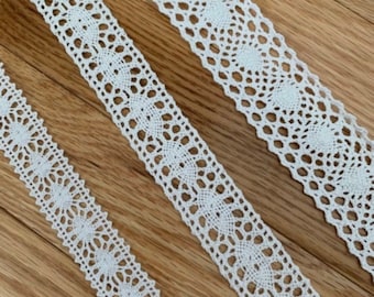 May include: Three white lace trims of varying widths, with a delicate floral pattern. The lace is arranged diagonally on a wooden surface.