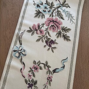 May include: A long, rectangular fabric runner featuring an off-white base with an intricate embroidered floral pattern. The design includes pink roses, light blue flowers, and purple wheat-like stalks, framed by a decorative green leaf border.