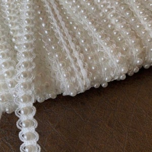 May include: White pearl trim with a scalloped edge. The trim is made of small white pearls sewn onto a white fabric base.