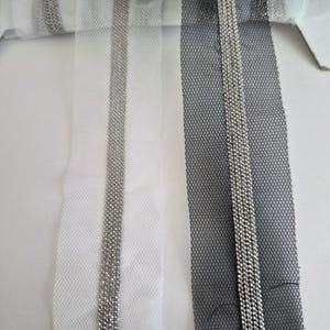 May include: Close-up of three rolls of decorative ribbon. One is black, one is white, and one is a light gray. Each ribbon has a mesh netting base with a vertical line of silver beads running down the center. The ribbons are on a white surface.