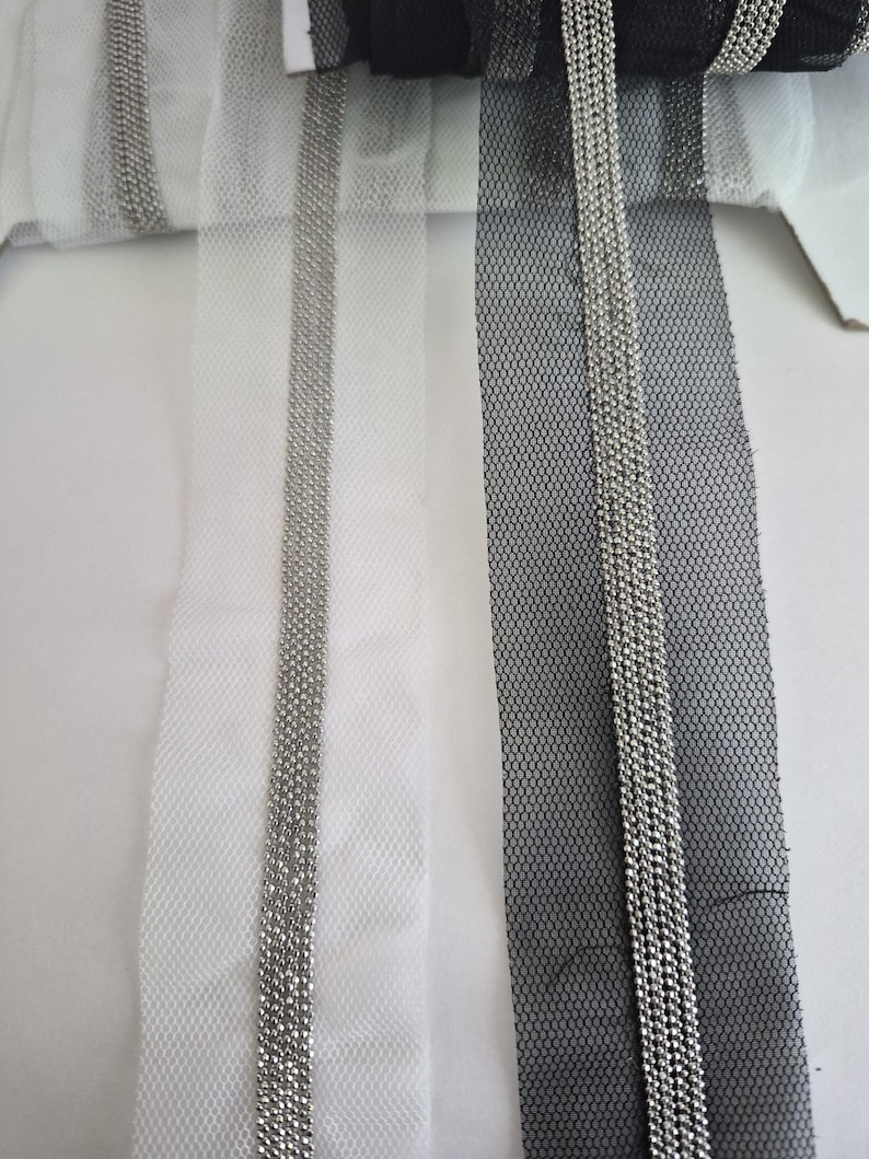 May include: Close-up of three rolls of decorative ribbon. One is black, one is white, and one is a light gray. Each ribbon has a mesh netting base with a vertical line of silver beads running down the center. The ribbons are on a white surface.