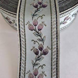 May include: A roll of decorative ribbon featuring a repeating floral pattern. The ribbon has a light blue background with a vertical design of purple flowers and green leaves. The edges are trimmed with black and gold accents.