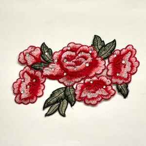 May include: Red embroidered flower applique with white accents and rhinestones. The applique features five flowers with green leaves.