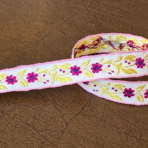May include: White ribbon trim with pink edges and yellow and pink floral embroidery.