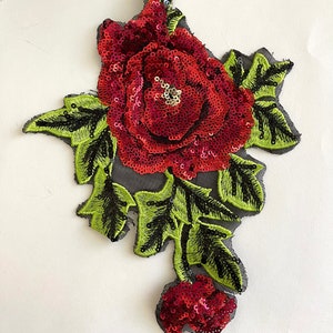 May include: A red sequined rose with green leaves and black stems on a black mesh background. The rose is a 3D design and has a shiny finish.