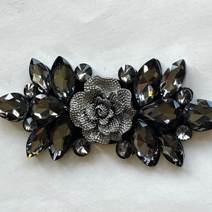 May include: A silver-toned flower embellishment with black rhinestone accents. The flower is surrounded by a cluster of black rhinestones.