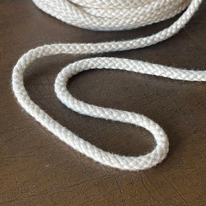 May include: A close-up of a white cotton cord, showing the braided texture. The cord is lying on a brown surface.