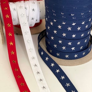 May include: Spools of ribbon in red, white, and navy blue, each adorned with star patterns. The red ribbon features gold stars, the white ribbon has silver stars, and the navy ribbon displays silver stars. Ideal for crafting and decorative projects.