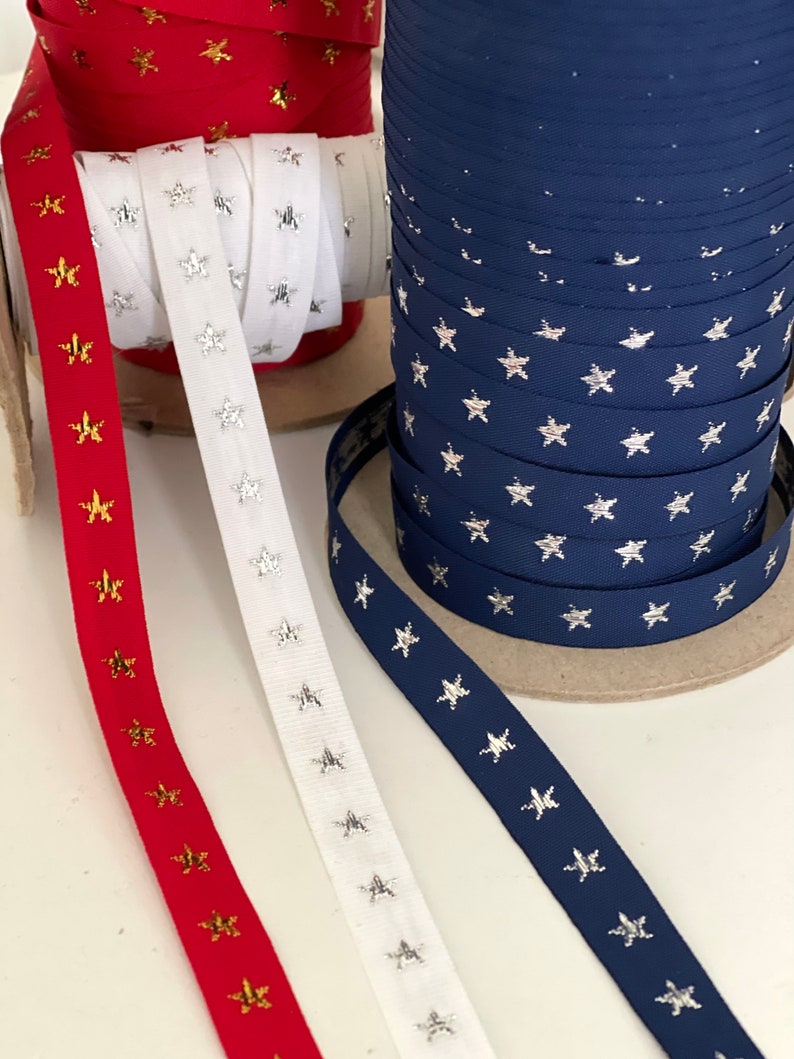 May include: Spools of ribbon in red, white, and navy blue, each adorned with star patterns. The red ribbon features gold stars, the white ribbon has silver stars, and the navy ribbon displays silver stars. Ideal for crafting and decorative projects.