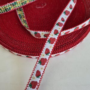 May include: A roll of red fabric with a textured surface. A white ribbon with a strawberry pattern and a scalloped red edge is visible. Another ribbon with a green, yellow, and white plaid pattern is also visible.