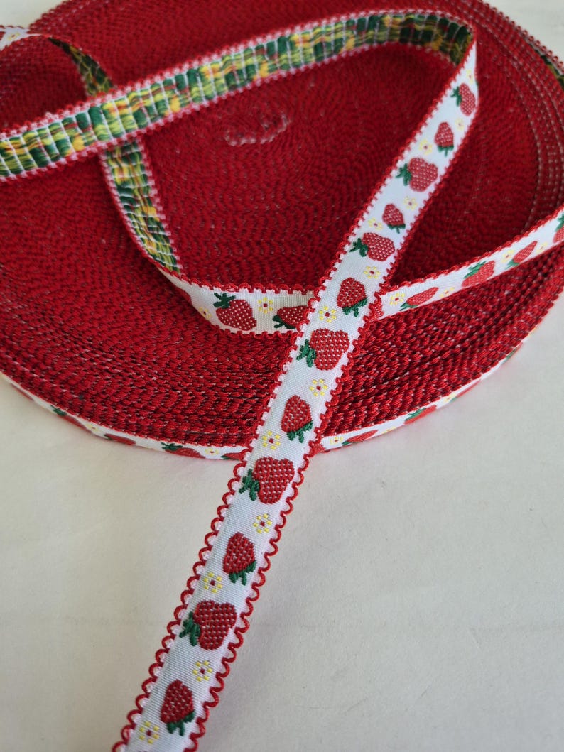May include: A roll of red fabric with a textured surface. A white ribbon with a strawberry pattern and a scalloped red edge is visible. Another ribbon with a green, yellow, and white plaid pattern is also visible.
