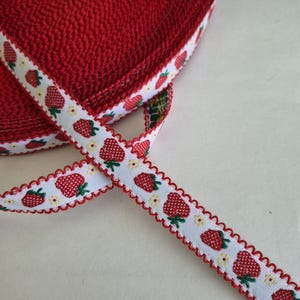 May include: A roll of red fabric with a white ribbon featuring a repeating pattern of red strawberries and yellow flowers. The ribbon has a scalloped edge, adding a decorative touch to the textile.