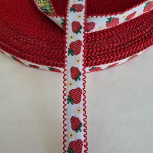 May include: A roll of red ribbon with a white border featuring a pattern of red strawberries and small yellow flowers. The ribbon's edge has a decorative, scalloped trim. The ribbon is likely used for crafts or gift wrapping.