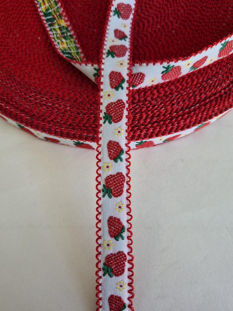 May include: A roll of red ribbon with a white border featuring a pattern of red strawberries and small yellow flowers. The ribbon's edge has a decorative, scalloped trim. The ribbon is likely used for crafts or gift wrapping.