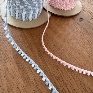 May include: Spools of decorative trim in pastel colors. One spool is light blue and white, the other is pink and white. A length of light blue and white trim and a length of pink and white trim are unspooled on a wooden surface.