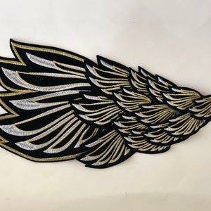 May include: A black and gold embroidered patch featuring a stylized wing design. The wing is detailed with multiple layers of feathers, creating a textured look.