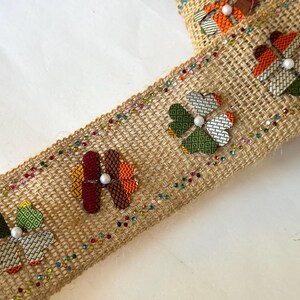 May include: A brown burlap ribbon with colorful fabric flowers and white pearl accents. The ribbon is decorated with a row of iridescent sequins.