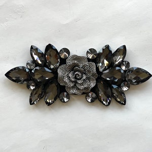 May include: A silver-toned flower embellishment with black rhinestone accents. The flower is surrounded by a cluster of black rhinestones.