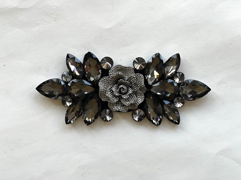 May include: A silver-toned flower embellishment with black rhinestone accents. The flower is surrounded by a cluster of black rhinestones.