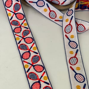 May include: White ribbon with a repeating pattern of red and blue tennis rackets and yellow tennis balls. The ribbon is folded in half, showing both sides of the pattern.