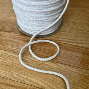 May include: A spool of white cotton rope, with a length of rope coiled around the spool and a loose end of rope extending from the spool.