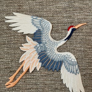 May include: Embroidered patch featuring a crane in flight. The patch showcases a detailed design with white, blue, and orange thread. The crane has a red head and a long, orange beak. The wings are spread wide, and the patch is likely for clothing or accessories.