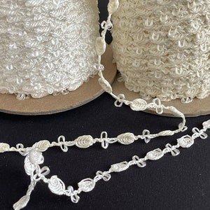 May include: Two spools of off-white decorative trim with a looped texture and a separate strand of the same trim. The trim features a repeating pattern of small, connected floral-like shapes. The spools are on cardboard bases.