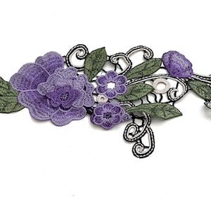 May include: Purple and green embroidered floral applique with black and silver thread details. The applique features a large rose and smaller flowers.