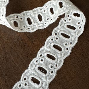 May include: Close-up of a white eyelet lace trim with a scalloped edge. The trim features a repeating pattern of oval cutouts and small floral details. The fabric is soft and delicate, suitable for various craft projects.