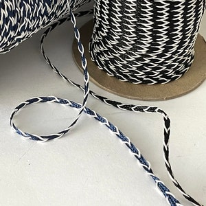 May include: A close-up of a black, white, and blue braided cord. The cord is wound around a cardboard spool. The cord is made of a thin, woven material.