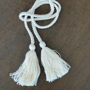 May include: A white braided rope with two tassels at the ends. The rope is tied in a loop and the tassels are hanging down.