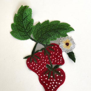 May include: Embroidered patch featuring three red strawberries with white seeds, a green stem, and a white flower with a yellow center.