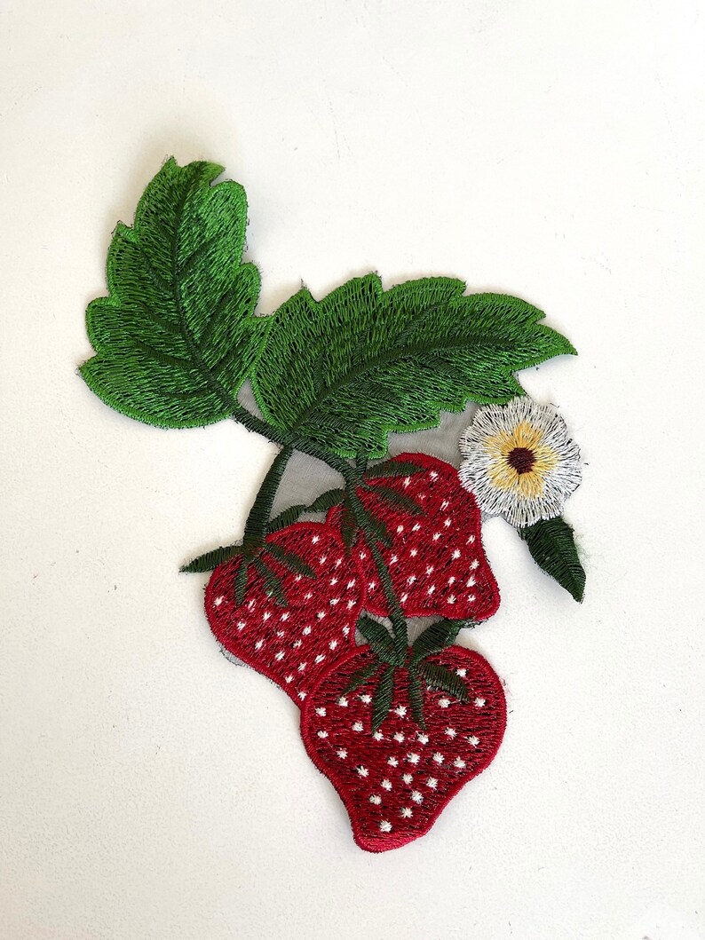 May include: Embroidered patch featuring three red strawberries with white seeds, a green stem, and a white flower with a yellow center.