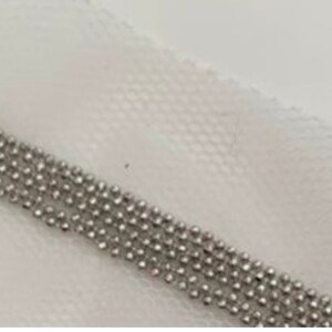 May include: Close-up of a silver beaded chain, composed of multiple strands of small, round beads. The chain is set against a white, textured fabric background, creating a contrast between the shiny metal and the soft textile.