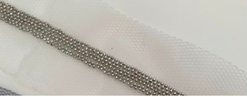 May include: Close-up of a silver beaded chain, composed of multiple strands of small, round beads. The chain is set against a white, textured fabric background, creating a contrast between the shiny metal and the soft textile.
