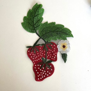 May include: Embroidered patch featuring three red strawberries with white seeds, green leaves, and a white flower with a yellow center.