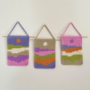 May include: Three crocheted wall hangings with abstract geometric designs in shades of green, purple, pink, orange, and brown. Each hanging has a white circle in the center.