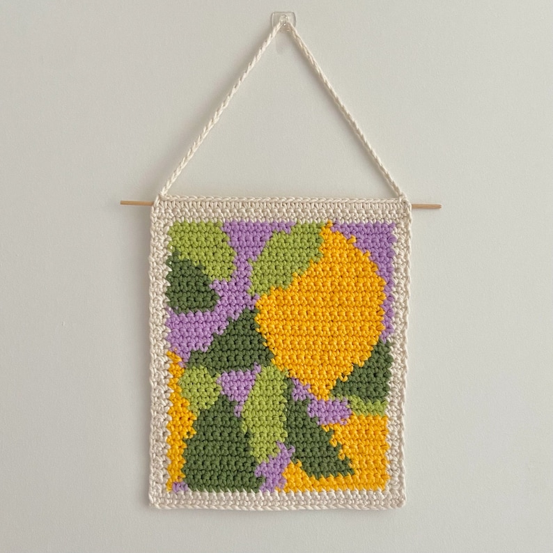 May include: A crocheted wall hanging with a geometric pattern in shades of green, yellow, and purple. The hanging is framed with a white crocheted border and is attached to a white wall with a string and a wooden dowel.