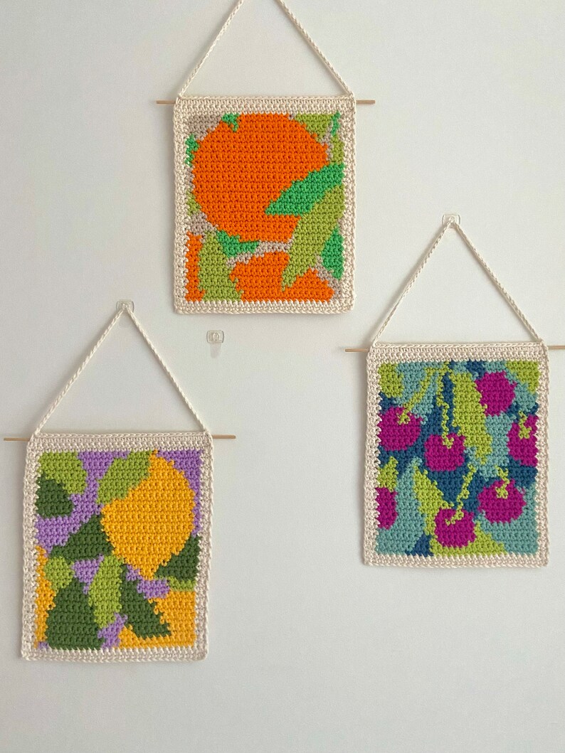 May include: Three crocheted wall hangings with geometric designs. The first features a large orange circle with green and white accents. The second features a yellow circle with green, purple, and white accents. The third features a cluster of pink circles with green, blue, and white accents.
