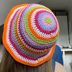 Pode incluir: Um chapéu de balde de crochê colorido com listras rosa, laranja, verde, branco e roxo.