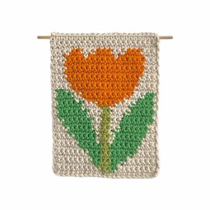 Puede incluir: Un tapiz de pared rectangular hecho a mano con ganchillo, con un vibrante diseño de tulipán naranja con hojas y tallo verdes sobre un fondo crema. La pieza está suspendida de una varilla de madera, mostrando un tema floral.