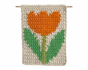 May include: A handmade, rectangular crochet wall hanging featuring a vibrant orange tulip design with green leaves and stem against a cream background. The piece is suspended from a wooden dowel, showcasing a floral theme.