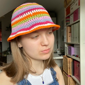 以下が含まれることがあります： オレンジ色のつばが付いた、カラフルなストライプのクロシェのバケットハットをかぶった人。帽子はピンク、グリーン、ホワイト、オレンジのストライプです。