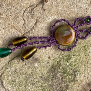 Puede incluir: Un collar de macramé morado con un colgante de cerámica marrón y verde y cuentas de vidrio marrón y verde.
