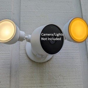 May include: White outdoor security light fixture with two white light bulbs and a black camera. The text "Camera/Lights Not Included" is printed on the camera.