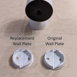 May include: Two white wall plates for a Google Nest Cam. The plate on the left is a replacement plate, and the plate on the right is the original plate.