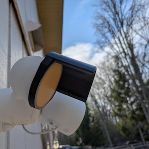 May include: A white security camera with a black sunshade mounted on a white wall. The camera is pointed towards the sky and the sunshade is blocking the sun from shining directly into the lens.